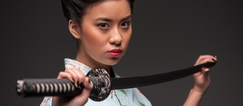 Total concentration. Side view portrait of young beautiful Japanese woman in kimono looking aggressively at camera and holding katana sword while standing against grey background